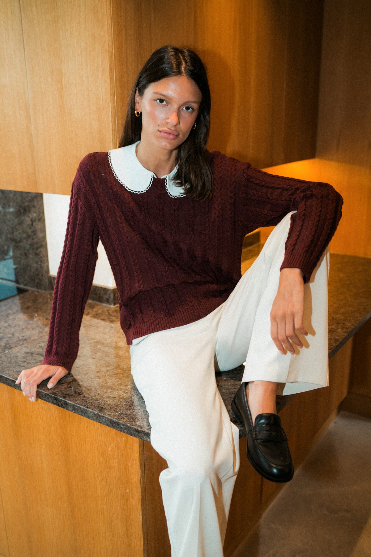 Girl sitting on counter of kitchen. She is wearing a red collared sweater, white trousers, and black loafers.