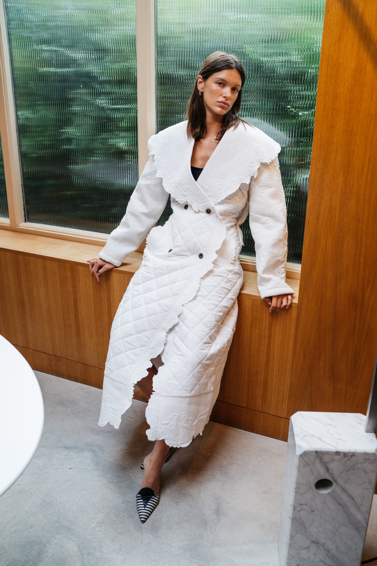 Girl leaning against a frosted glass window, she is wearing an ivory quilted trench coat.
