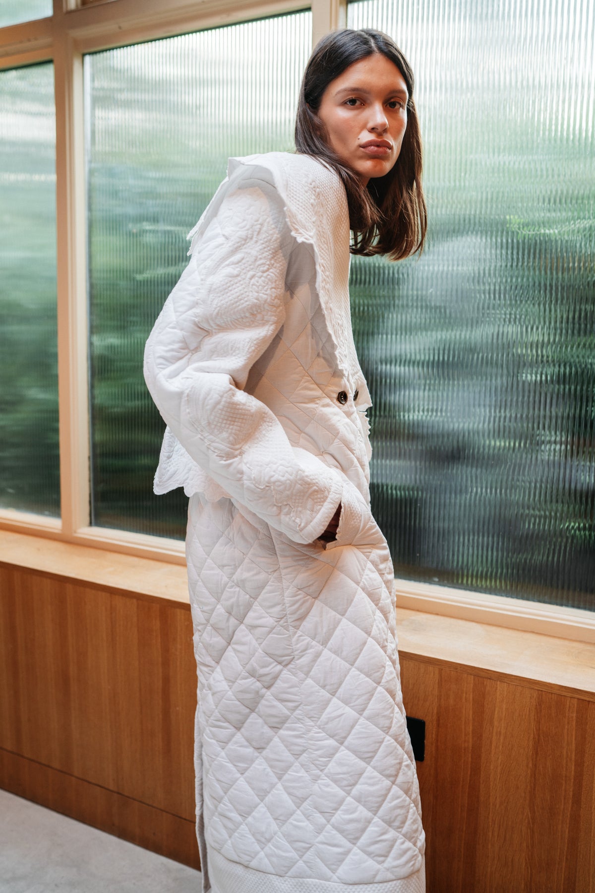 Girl standing in front of frosted glass, facing to the side, showcasing side sleeve detail of quilted trench coat.