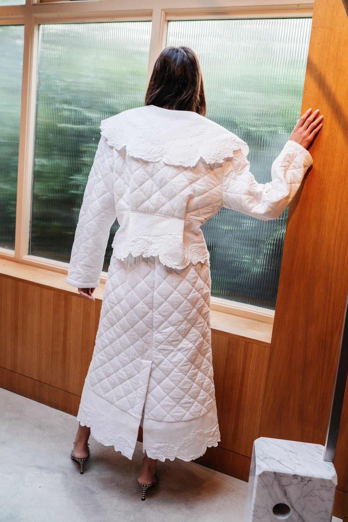 Girl standing in front of frosted windows, facing away from the camera. She wears a quilted trench coat.