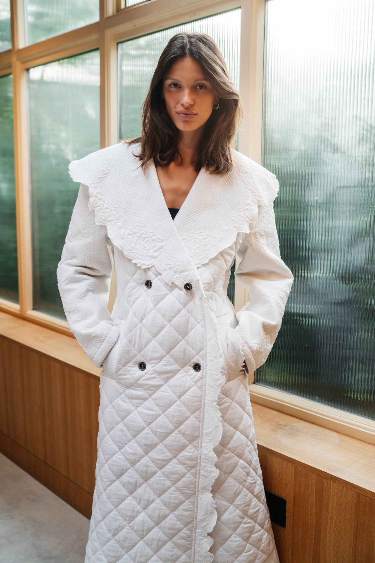 Girl standing in front of frosted glass, wearing ivory quilted trench coat.
