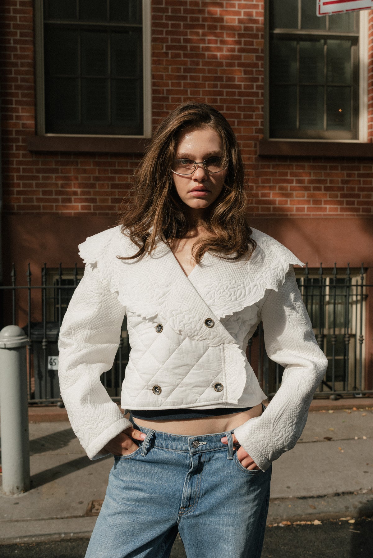 Girl standing outside, the sun shines across her. She has her hands tucked in her blue denim jean pockets and is wearing an ivory quilted coat.