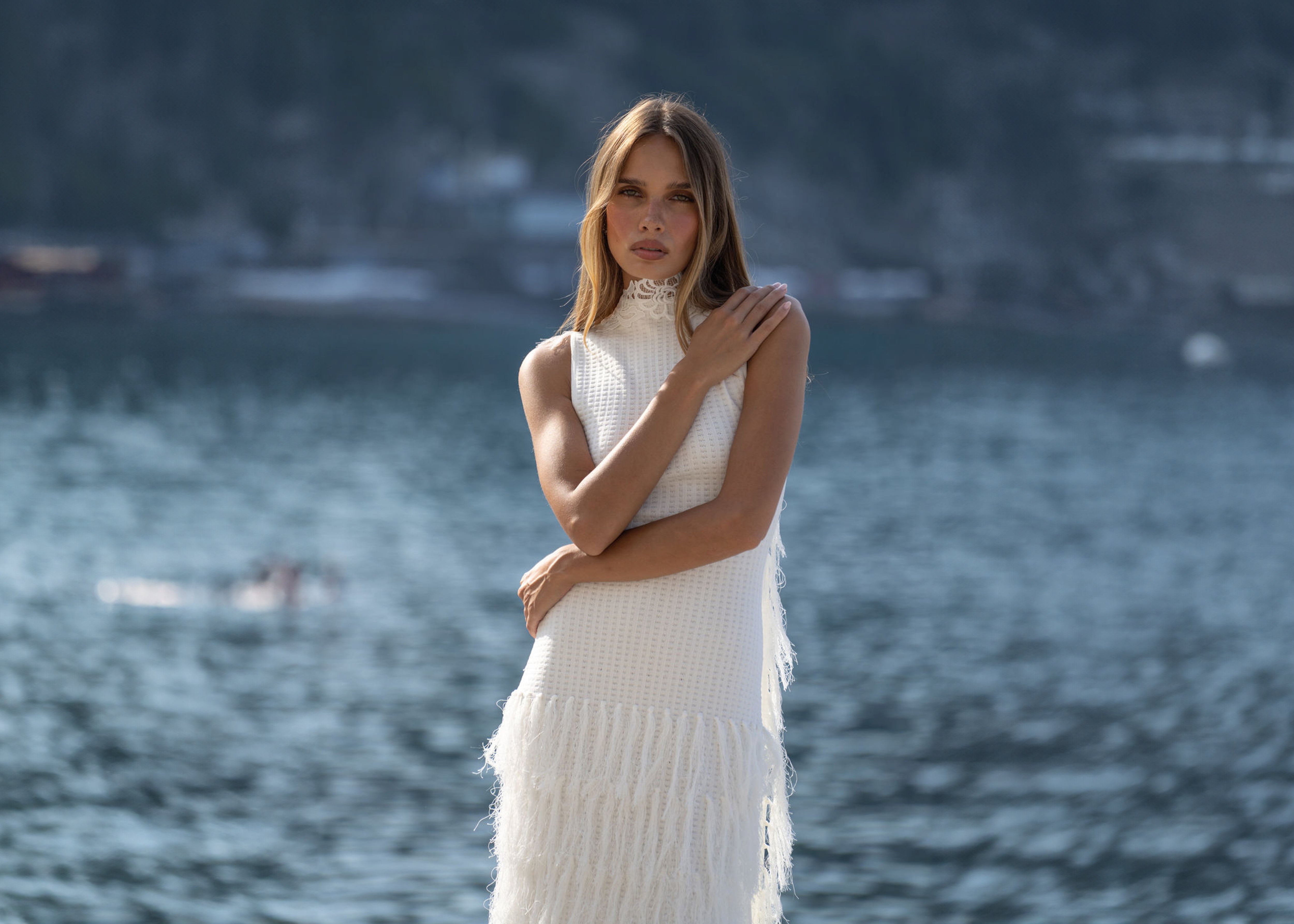 Woman in a white dress standing by a body of water with mountains in the background