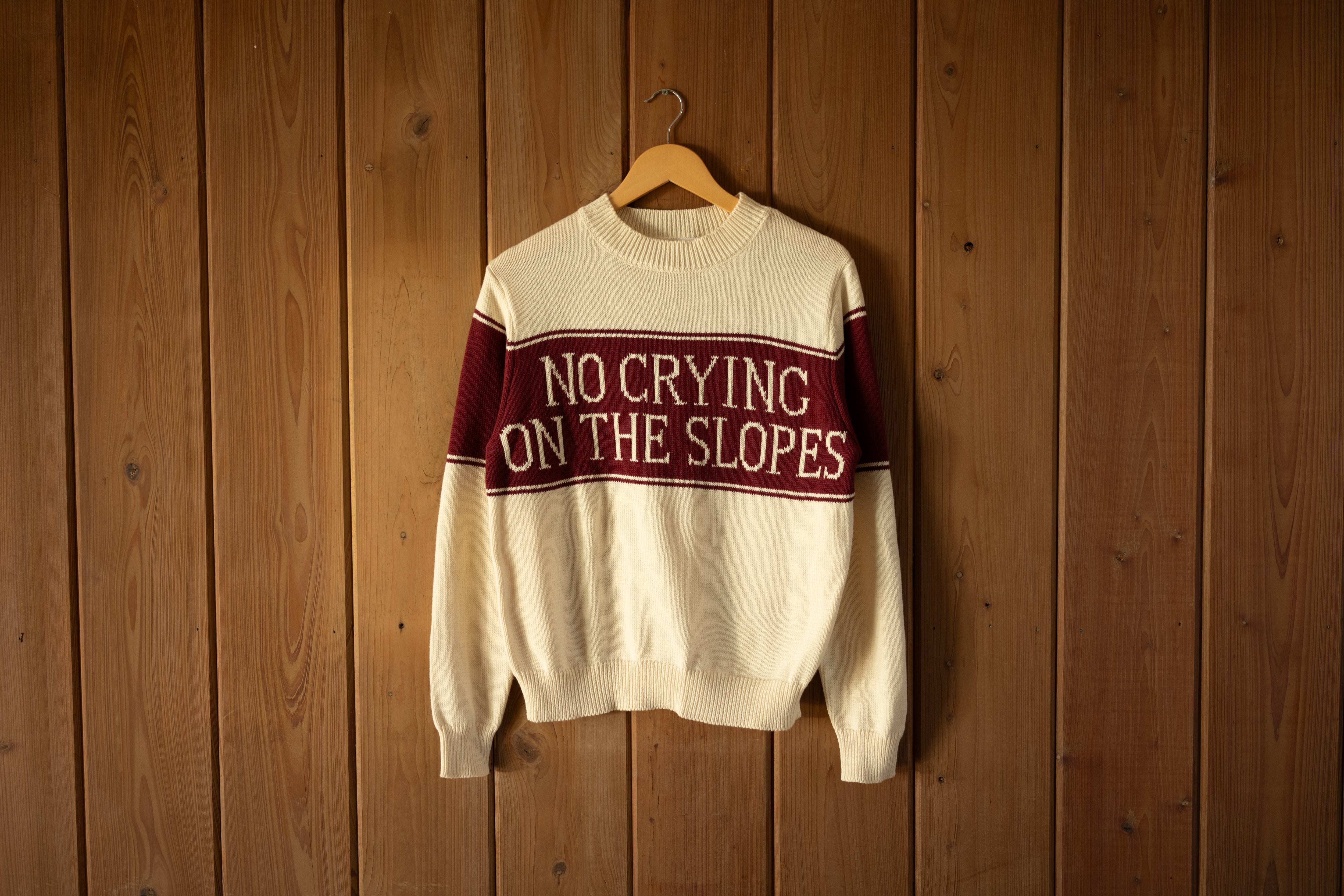 Sweater with 'No Crying on the Slopes' text hanging on a wooden wall
