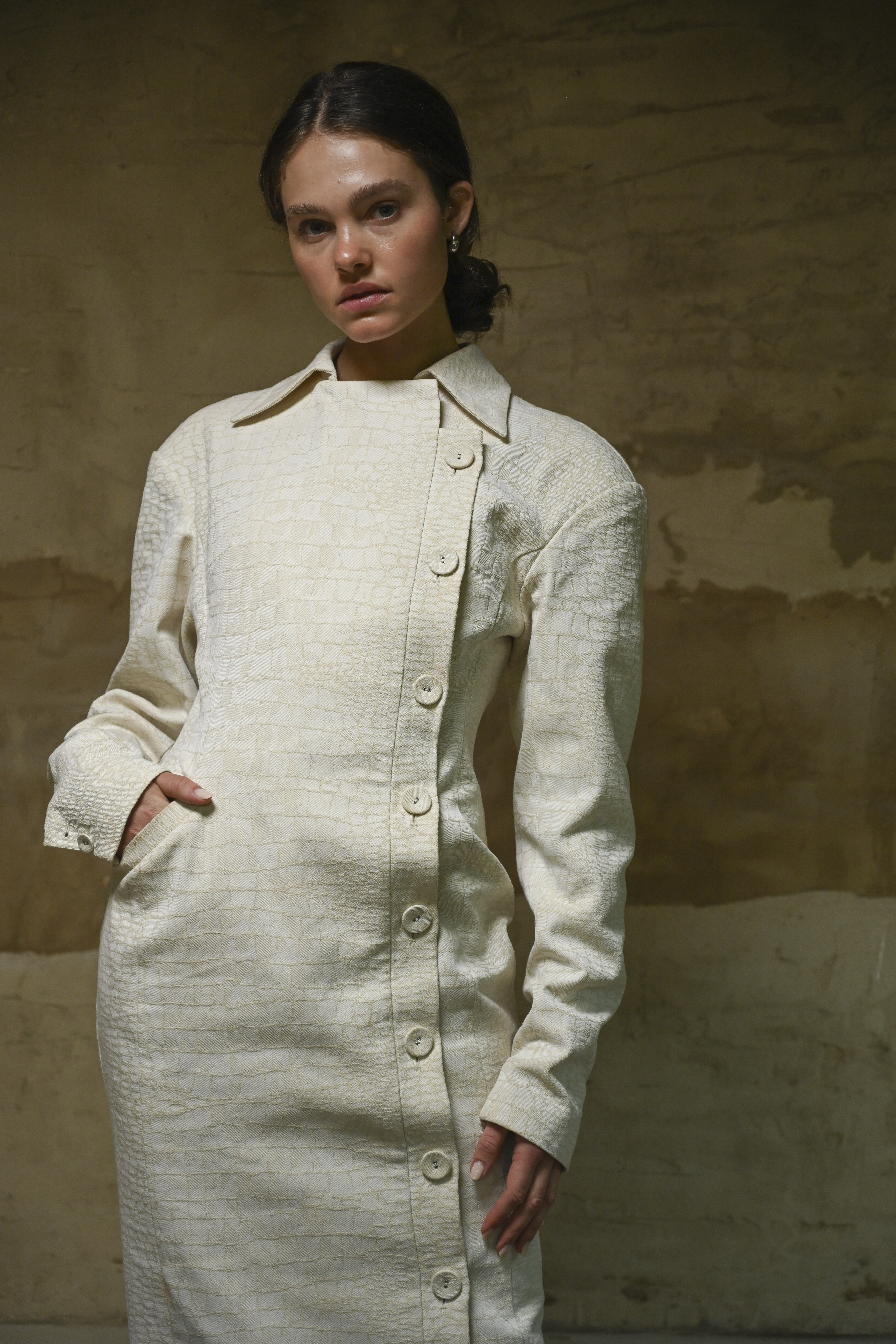 Woman wearing a long, light-colored coat against a textured beige wall.