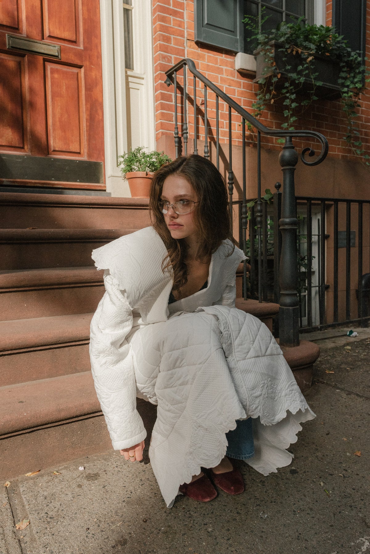Girl sitting on the steps of a brownstone home, she looks off to her right. She is wearing an ivory quilted trench coat. 