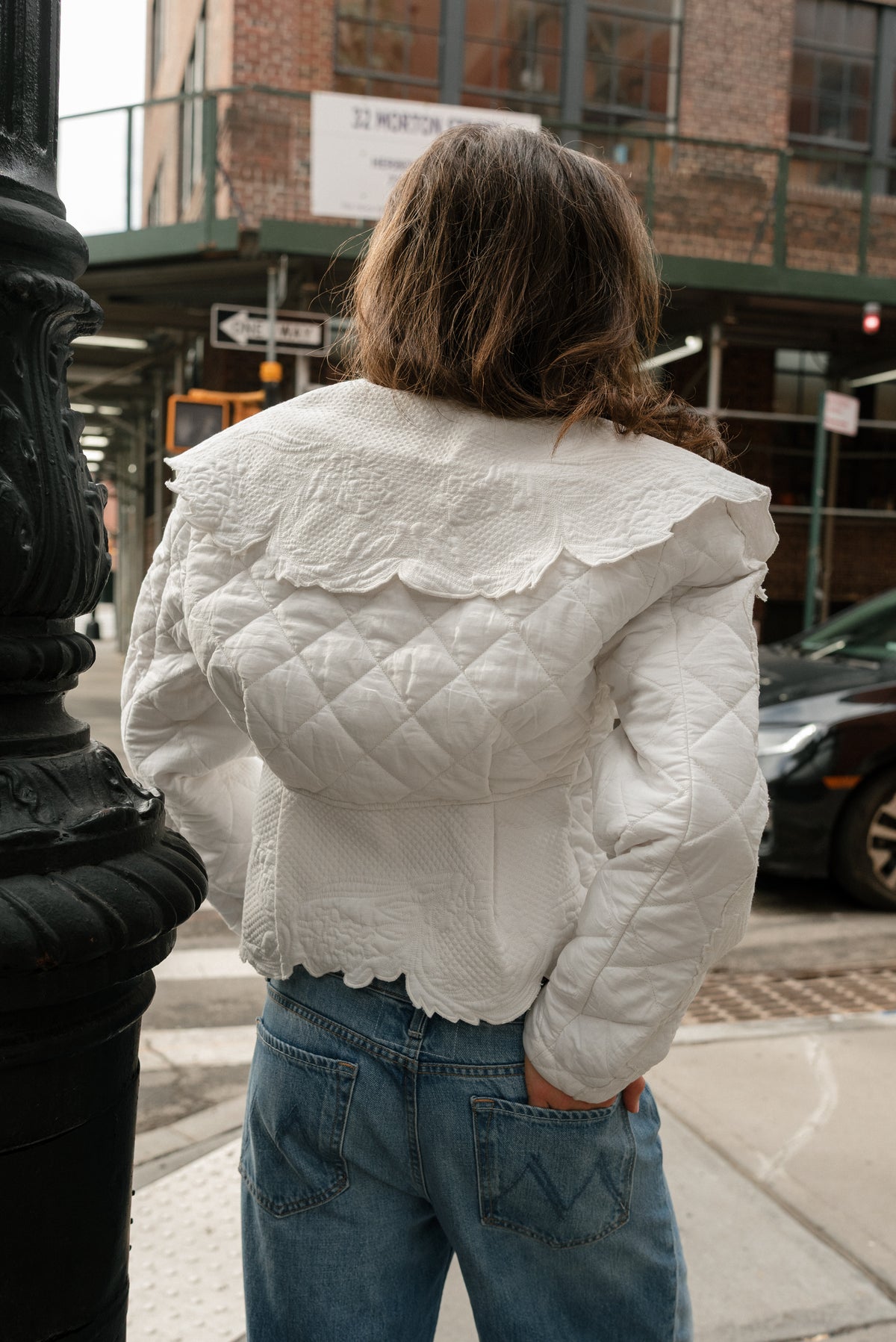 Girl is facing away from the camera, showcasing the back of the ivory cropped quilted coat she is wearing. Her hand is in the back pocket of her blue jeans.