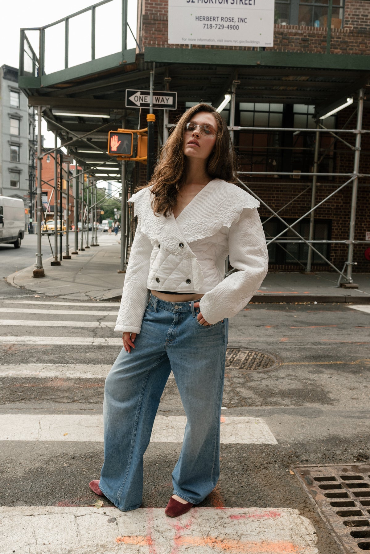 Girl standing on a crosswalk, she leans back with one hand in her pocket. She wears a pair of blue jeans and an ivory quilted jacket. 