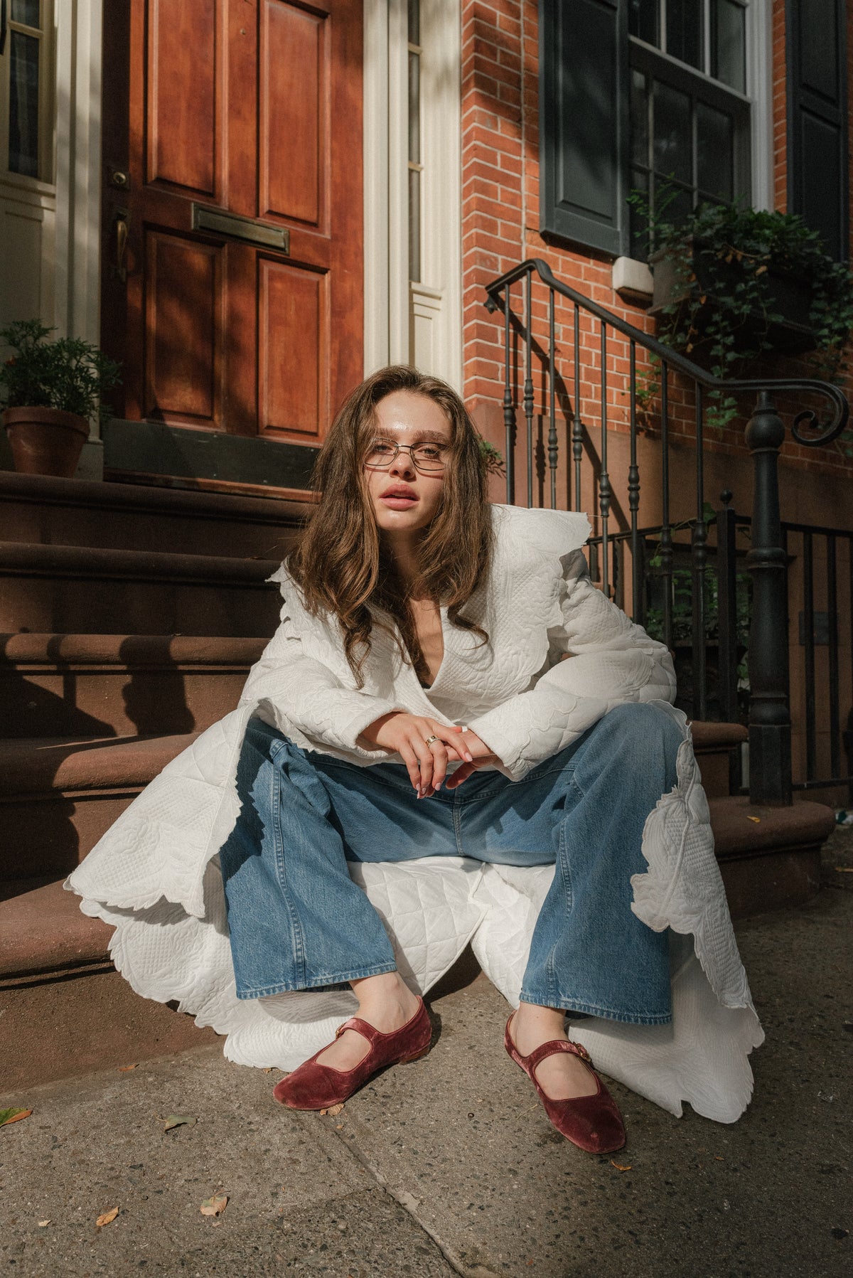 Girl sitting on steps of brownstone home outdoors, leaning forward toward camera. Wearing a quilted trench coat. 